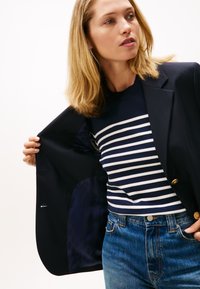 Mornarski blazer z zlatimi gumbi, nošen čez mornarsko in belo črtasto pulover, v kombinaciji z modrimi kavbojkami. Notranjost ima mornarsko vzorčasto podlogo.
