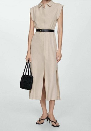 Beiges ärmelloses Midikleid mit Kragen und vorderen Knöpfen, schwarzer Gürtel, schwarze gewebte Handtasche und schwarze Sandalen mit Absatz.