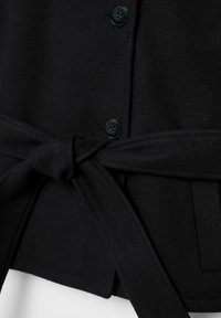 Primo piano di un cappotto di lana nero che mostra due bottoni neri e una cintura di tessuto annodata intorno alla vita.