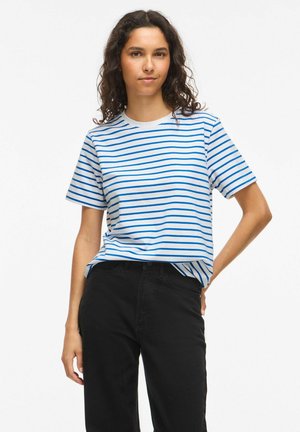 Femme aux cheveux bouclés portant un t-shirt blanc à rayures bleues et un pantalon noir, debout avec une main sur la hanche.
