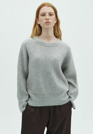 Jeune femme aux longs cheveux roux portant un pull ample gris clair côtelé et un pantalon foncé, debout devant un fond blanc uni.