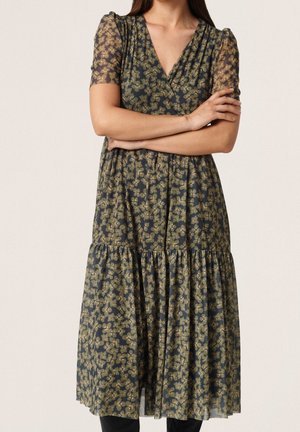 Femme portant une robe vert foncé à imprimé floral jaune, manches courtes bouffantes et jupe froncée, debout les bras croisés.