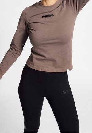 Osoba ubrana w brązową koszulę z długim rękawem oraz czarne legginsy z logo "Hummel", stojąca z uniesioną jedną ręką na jednolitym tle.