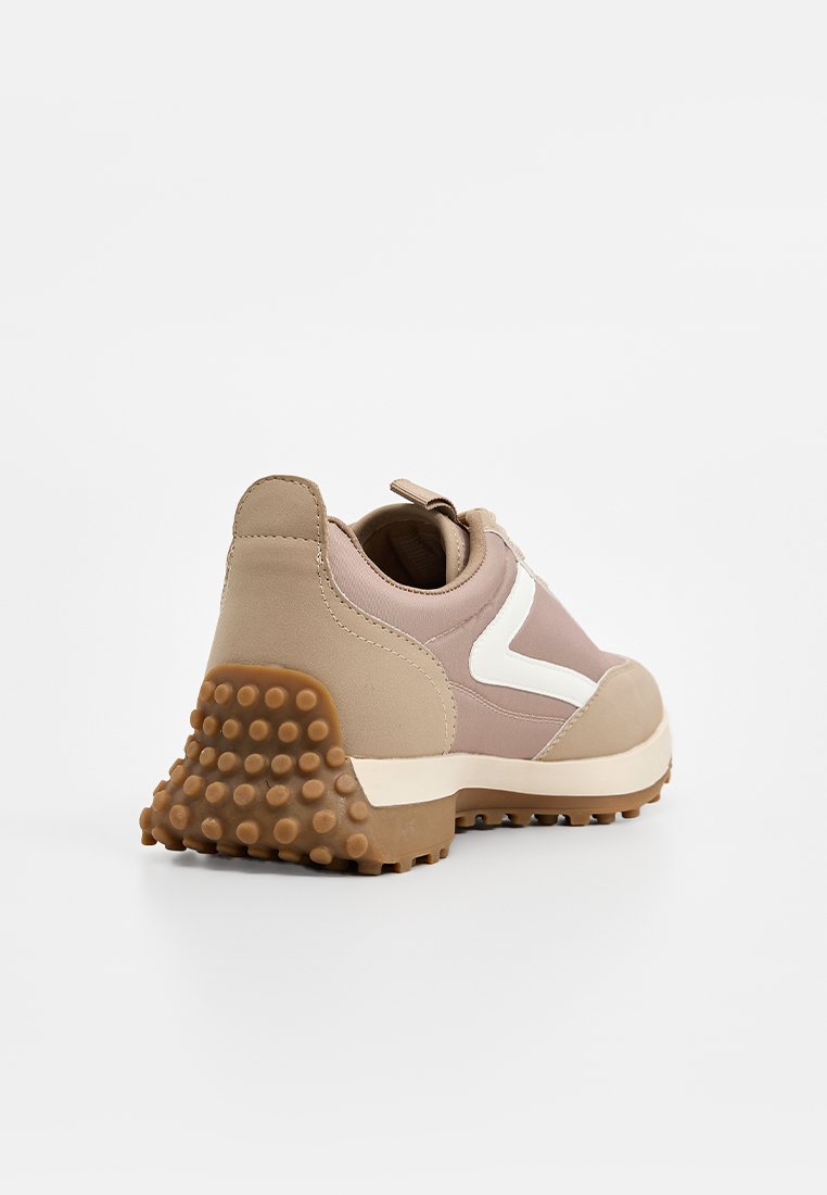 Zapatilla deportiva beige y rosa con una suela de goma texturizada, forma redondeada y un acento blanco. Presenta un acabado de tela en la parte superior y un talón acolchado.