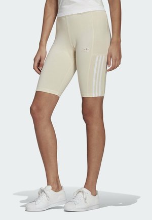 Kvinde iført beige Adidas cykelshorts til knælængde med hvidt logo og striber, kombineret med hvide sneakers og hvid top.