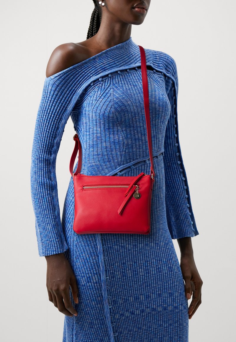 Borsa a tracolla in pelle rossa con tasca zip e dettaglio in frangia, indossata sopra un vestito azzurro a costine con design off-shoulder.