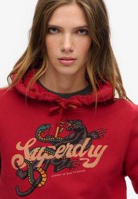 Sudadera roja con capucha que presenta una impresión gráfica de un dragón y la palabra "Superdry" en dorado, con capucha ajustable y cuello acanalado.