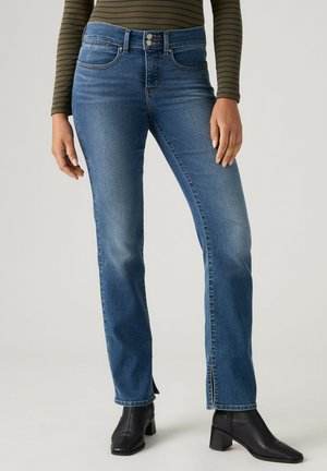 Person trägt mittelhohe blaue Jeans mit leichtem Schlag und vorderen Schlitzen kombiniert mit schwarzen Stiefeletten und einem grün gestreiften Langarmshirt.