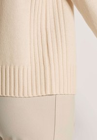 Nahaufnahme des Saums eines beigen gerippten Strickpullovers, kombiniert mit hellbeigen strukturierten Hosen, die die Stofftexturen und Nahtdetails hervorhebt.