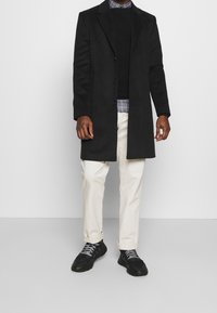 Cappotto lungo nero sopra un maglione nero, camicia a quadri, pantaloni chiari e sneakers nere con lacci grigi. Tessuto liscio, vestibilità sartoriale.