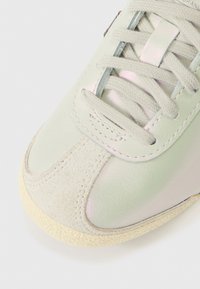 Ténis verde claro com parte superior em couro liso, biqueira em camurça e atacadores planos. Apresenta um brilho subtil e sola de borracha texturizada.