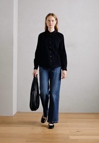 Chemise noire à boutons, jeans bleus à large jambe, chaussures noires et sac noir. Textures de tissu lisses sur un fond neutre et un sol en bois.