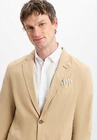 Blazer beige leggero con rever a richiesta, chiusura con un bottone e una tasca sul petto contenente un fazzoletto a righe. Camicia bianca sotto.