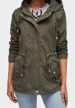 Nainen pukeutuneena oliivinvihreään hupulliseen takkiin, jossa on säädettävä vyötärönauha ja etutaskut läpällä, yhdistettynä vaaleansinisiin farkkuihin ja raitapaitaan.