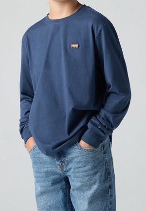 Person trägt ein dunkelblaues Levi's Langarmshirt mit kleinem Logo-Patch auf der Brust, Hände in den Taschen der hellblauen Jeans.
