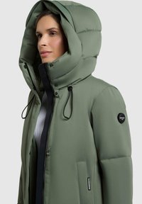 Giacca puffer verde oliva con un grande cappuccio, zip frontale, cordini regolabili e una patch con logo sulla manica sinistra. Presenta una texture liscia.