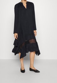 Černé plisované šaty s dlouhými rukávy, výstřihem do V, asymetrickým lemem, volánkovými akcenty a průhlednou spodní částí. V kombinaci s černými balerínami.