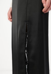 Svart satengskjørt med sidelitsover og eksponert glidelås med merketrekksnor som viser "KAR LAGERFELD JEANS." Glatt tekstur.