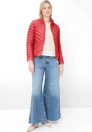 Frau trägt eine rote gesteppte Jacke, cremefarbenen, knöpfbaren Cardigan, hellblaue Jeans mit weitem Bein und cremefarbene flache Schuhe, steht vor weißem Hintergrund.