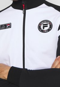 Giacca bianca con zip e maniche nere. Presenta il logo FILA ricamato e il testo "fai le tue regole", con una texture liscia e un design aderente.
