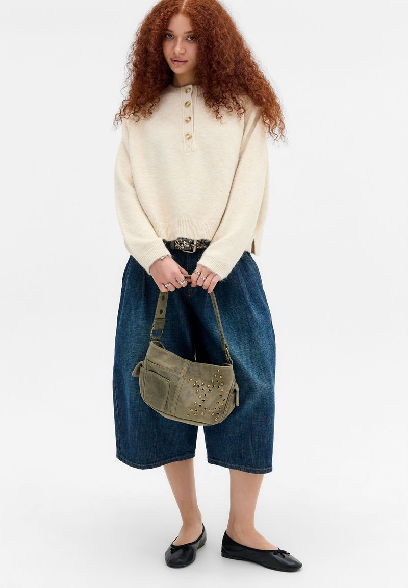 Jeune femme aux cheveux roux bouclés portant un pull beige, un pantalon large en jean, des ballerines noires, tenant un sac à main vert clouté.