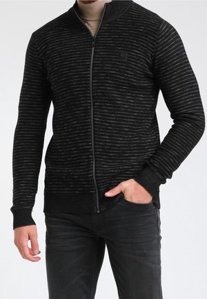Homme portant un pull zippé rayé noir et gris sur un col roulé beige, une main dans la poche d'un jean noir.