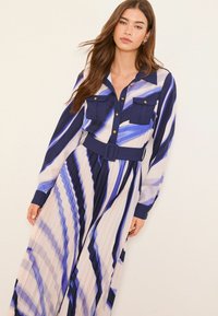 Langes, plissiertes Kleid in Blau- und Cremetönen. Verfügt über eine geknöpfte Vorderseite, zwei Brusttaschen und einen Stoffgürtel. Glattes, leichtes Material.