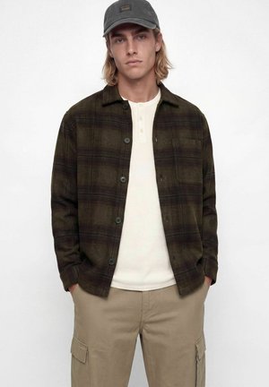 Jeune homme portant une chemise à carreaux foncés boutonnée sur un henley blanc, un pantalon cargo kaki et une casquette de baseball grise sur un fond uni.