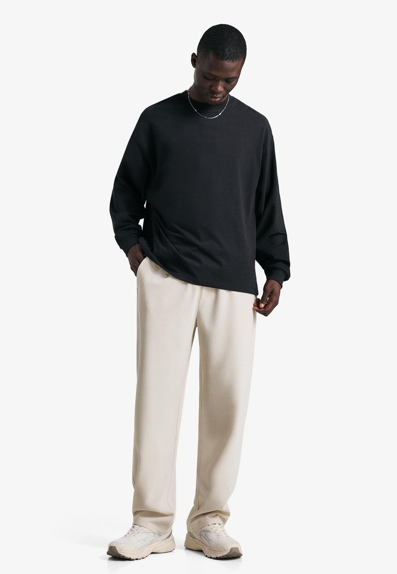 Cămașă neagră cu mâneci lungi și guler rotund, asortată cu pantaloni bej, lejeri. Modelul stă cu mâinile în buzunare, purtând pantofi sport de culoare deschisă.