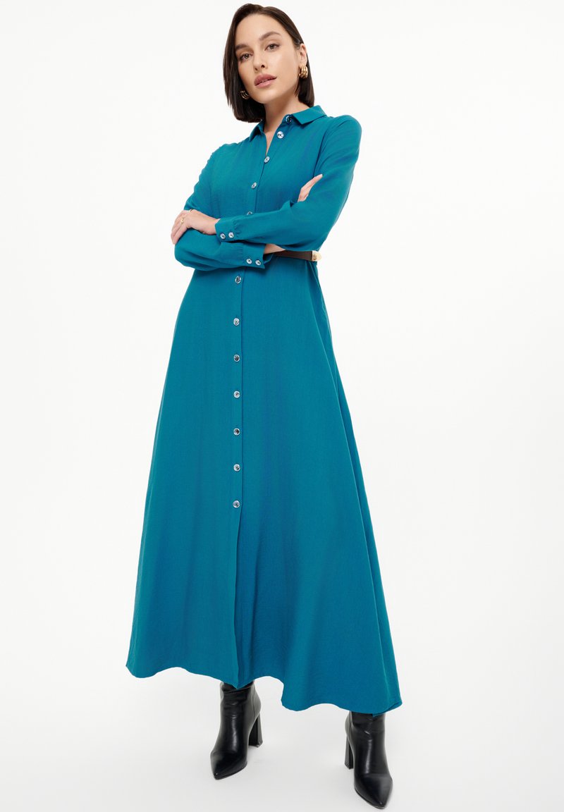 Abito lungo teal realizzato in tessuto leggero, con chiusura frontale a bottoni, maniche lunghe e una gonna fluida. Stivali neri con tacco ne completano il look.