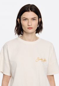 Lichtbeige katoenen T-shirt met een relaxte pasvorm. Heeft korte mouwen en een klein gouden geborduurd logo aan de linkerkant.