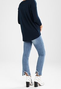 Marineblauwe lange mouw top met een relaxte pasvorm, gecombineerd met lichtblauwe jeans met rafelige uiteinden en witte enkellaarsjes met zwarte blokhakken.