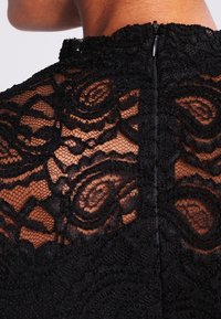 Sort blondekjole med høj halsudskæring, der har et blomstermønster, gennemsigtig tekstur og lynlås bagpå. Detaljeorienteret design.