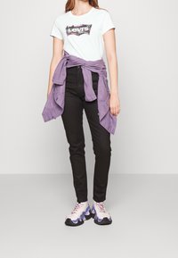 Biała koszulka Levi's z kolorowym logo, czarne obcisłe jeansy oraz lawendowa, oversize'owa koszula związana w talii. Grube białe i fioletowe sneakersy.