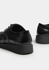 Une paire de chaussures brogues en cuir verni noir avec des semelles en caoutchouc noir épaisses, sur un fond gris clair, montrant des vues de dos et de côté.