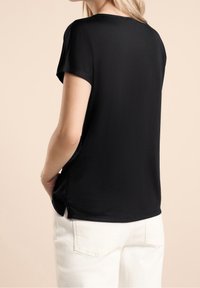 Svart t-shirt med kort ärm och rund hals, mjuk textur och sidofickor, ihop med ljusa byxor. Enkelt design och passform.