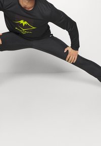 Mustat pitkähihaiset urheilupaidat, jossa on vihreä vuoristologo ja teksti. Kiinnitetyt mustat leggingsit. Malli suorittaa haarahypyn venytyksen.