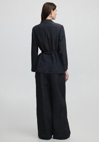 Completo nero con texture, maniche lunghe e vita con cintura. I pantaloni a gamba larga completano la giacca, creando un design minimalista.