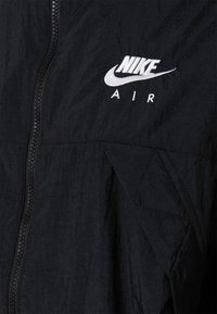 Svart vindjacka med dragkedja och en texturerad yta. Broderad vit "Nike Air"-logotyp på övre vänstra sidan.