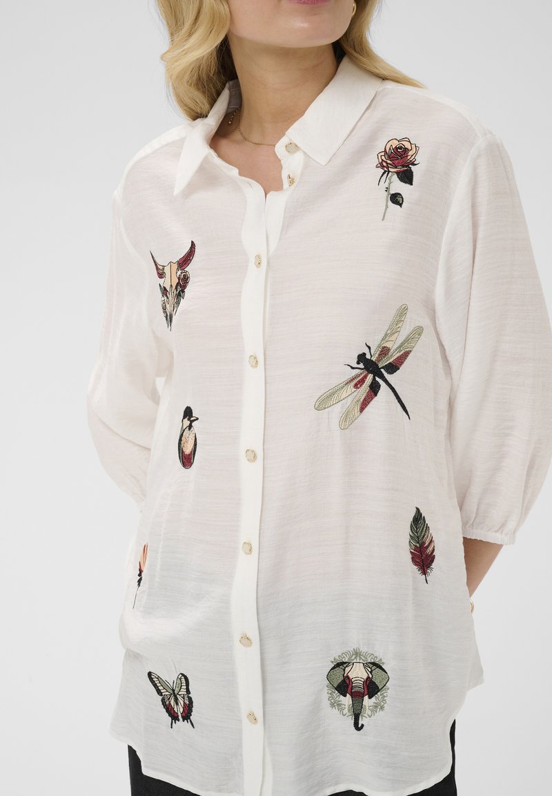 Femme portant une chemise blanche à boutons avec des motifs brodés de libellule, rose, papillon, éléphant, plume et crâne d'animal.