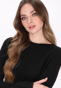 Pulover negru tricotat, cu o suprafață texturată și un decolteu larg. Modelul are părul lung și ondulat, machiaj minimal și trăsături ușor definite.