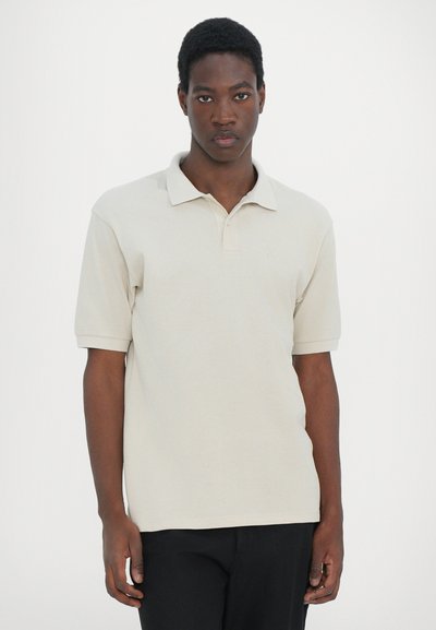 Polo beige à manches courtes, col classique, en coton doux, avec un détail logo subtil sur la poitrine, associé à un pantalon sombre.