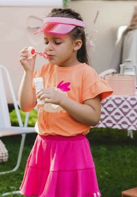 Kind dat een oranje shirt met een grafisch ontwerp en frilly mouwen draagt, gecombineerd met een roze, gelaagd rokje, terwijl het bellen blaast met een toverstok.