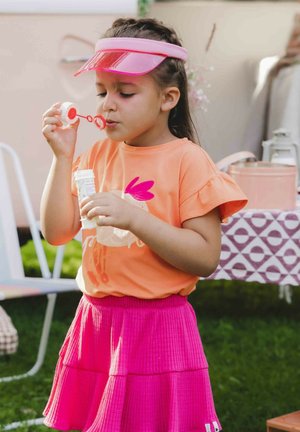 Kind dat een oranje shirt met een grafisch ontwerp en frilly mouwen draagt, gecombineerd met een roze, gelaagd rokje, terwijl het bellen blaast met een toverstok.