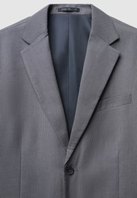 Grau gemusterter Blazer mit Reverskragen, Einzelknopfverschluss und einer Brusttasche. Innenfutter aus einfarbigem dunklem Material. Größenetikett sichtbar.