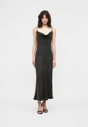 Femme aux cheveux bouclés portant une longue robe nuisette noire à fines bretelles et des sandales à talons hauts noires, debout devant un fond uni.