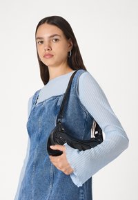 Geantă din piele neagră tip crossbody cu o formă curbată, închidere cu fermoar și curea ajustabilă. Modelul poartă un top cu nervuri albastre și o rochie din denim.