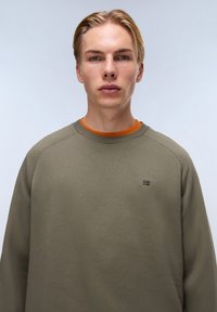 Olijfgroene sweatshirt met ronde hals, voorzien van een geribde kraag en manchetten, met een oranje binnenlaag en een klein logo detail op de borst.