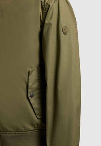 Manche de veste vert olive avec une poche à bouton-pression et un logo circulaire en patch sur le haut du bras, sur un fond gris clair.
