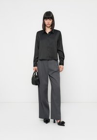Camisa de seda preta de botões com mangas compridas, combinada com calças cinza de cintura alta. A modelo segura uma pequena bolsa preta texturizada. Sapatos de salto pretos.